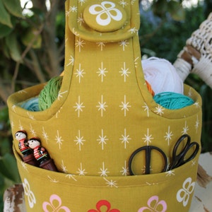 May include: A mustard yellow fabric yarn tote bag with a white star pattern. The bag has a handle and a pocket on the front. The pocket has two wooden figures and a pair of scissors inside. The bottom of the bag has a white, pink, and red flower pattern.