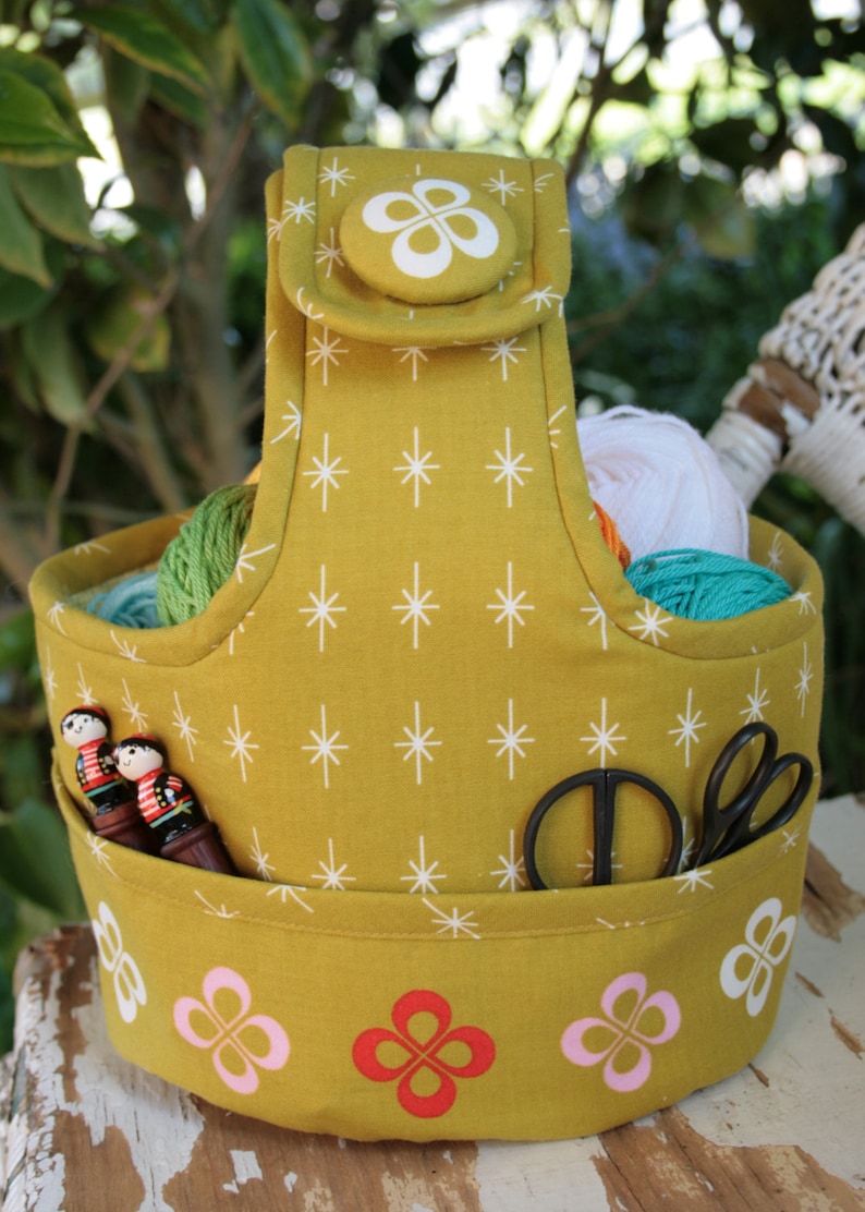 May include: A mustard yellow fabric yarn tote bag with a white star pattern. The bag has a handle and a pocket on the front. The pocket has two wooden figures and a pair of scissors inside. The bottom of the bag has a white, pink, and red flower pattern.