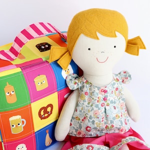 Puede incluir: Una mu&ntilde;eca hecha a mano con cabello amarillo, un vestido floral y una falda rosa. La mu&ntilde;eca est&aacute; sentada junto a una bolsa de lona de retazos colorida con varias ilustraciones de comida.