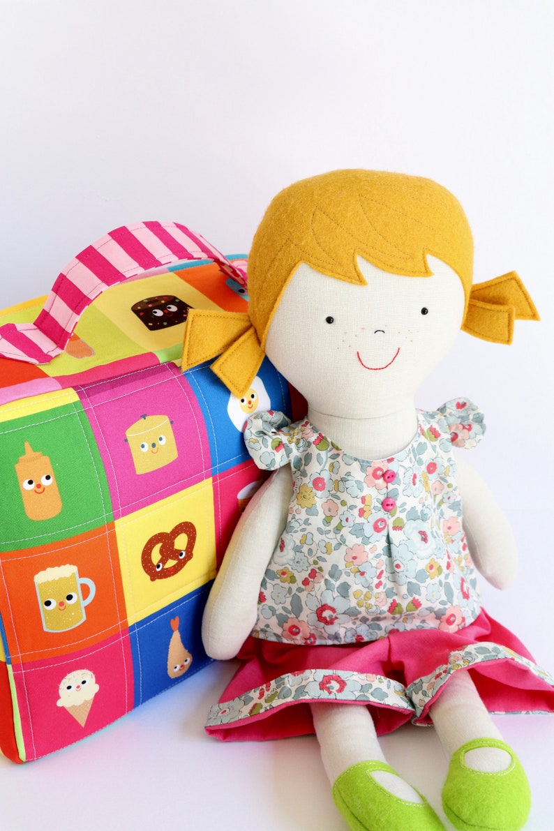 Puede incluir: Una mu&ntilde;eca hecha a mano con cabello amarillo, un vestido floral y una falda rosa. La mu&ntilde;eca est&aacute; sentada junto a una bolsa de lona de retazos colorida con varias ilustraciones de comida.