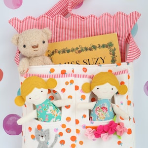 May include: A white fabric tote bag with orange and pink polka dots. The bag has two pockets with doll-sized openings. Two dolls with blonde hair and floral dresses are peeking out of the pockets. A teddy bear and a book titled "Miss Suzy" are inside the bag.