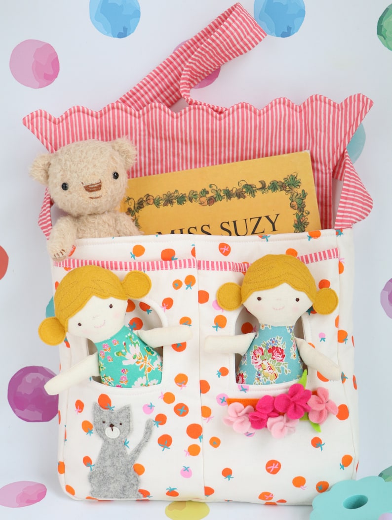 May include: A white fabric tote bag with orange and pink polka dots. The bag has two pockets with doll-sized openings. Two dolls with blonde hair and floral dresses are peeking out of the pockets. A teddy bear and a book titled "Miss Suzy" are inside the bag.