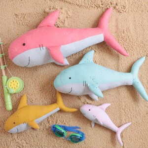 May include: Four plush shark toys in different colors and sizes. A pink shark, a light blue shark, a yellow shark, and a pink shark. The sharks are lying on a sandy surface.