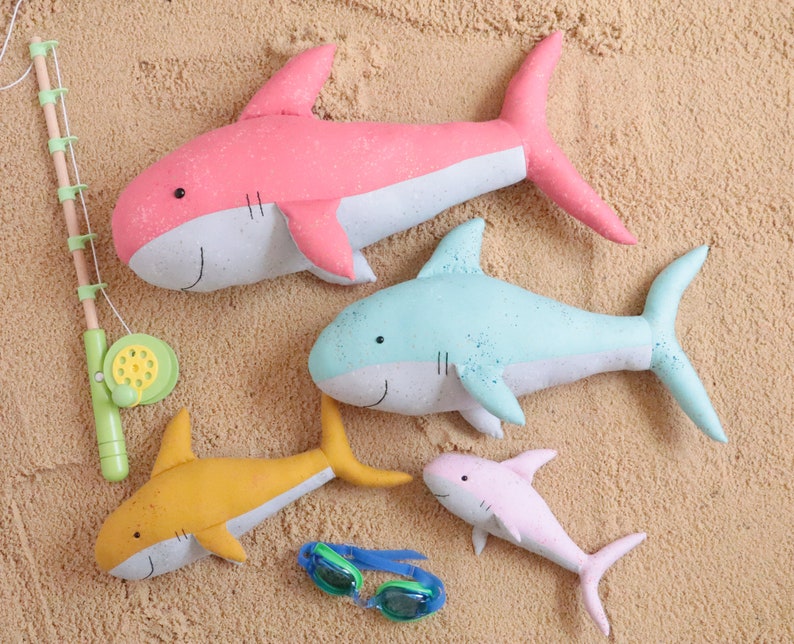 May include: Four plush shark toys in different colors and sizes. A pink shark, a light blue shark, a yellow shark, and a pink shark. The sharks are lying on a sandy surface.