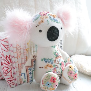 May include: A white plush koala bear with pink fur ears, embroidered with colorful flowers and sitting on a white wooden surface.
