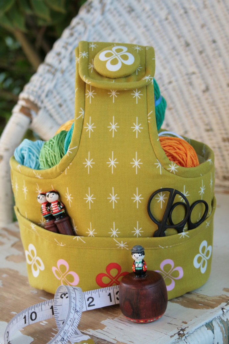 May include: A yellow fabric storage basket with a white star pattern and a white clover pattern. The basket has a handle and is filled with yarn balls, wooden figures, and a pair of scissors. A measuring tape is in the foreground.