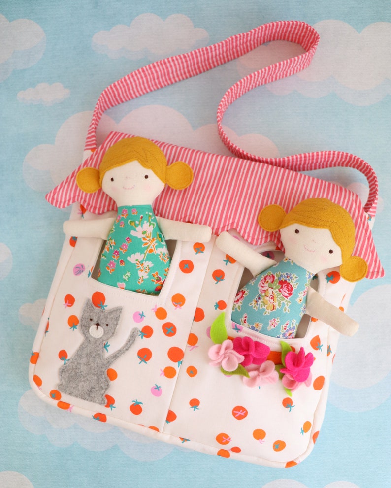 May include: A white fabric bag with a pink and white striped strap. The bag has multiple pockets and features a felt cat, felt flowers, and two felt dolls with yellow hair and floral dresses.