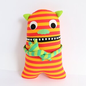May include: A striped plush toy monster with red and yellow stripes, green arms, and white eyes. The monster has a green nose and a black and white beaded necklace.
