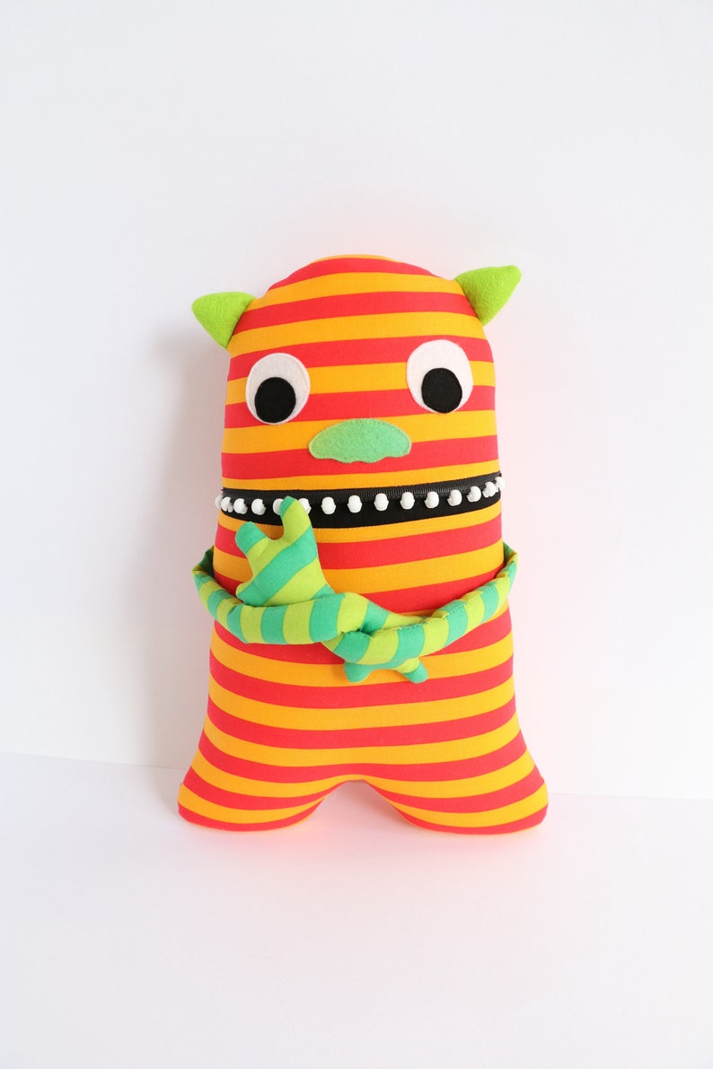 May include: A striped plush toy monster with red and yellow stripes, green arms, and white eyes. The monster has a green nose and a black and white beaded necklace.