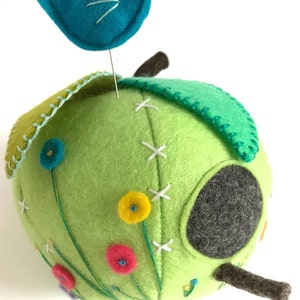 May include: A green felt birdhouse with a small round opening, decorated with embroidered flowers and a blue felt bird perched on top.