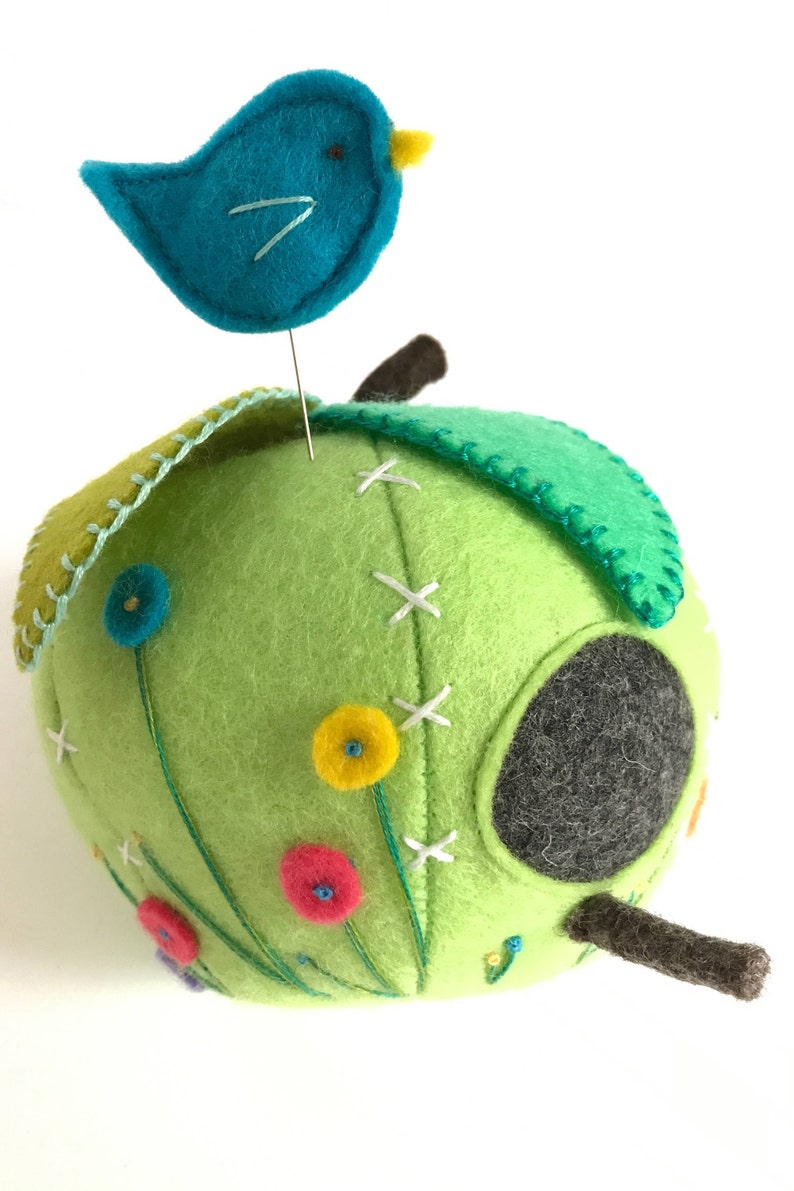 May include: A green felt birdhouse with a small round opening, decorated with embroidered flowers and a blue felt bird perched on top.