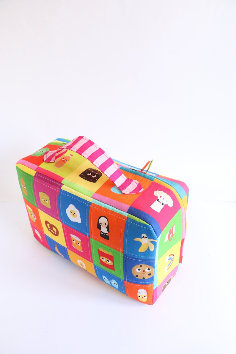 Puede incluir: Una caja de almacenamiento de tela colorida con un asa a rayas rosa y blanca. La caja est&aacute; decorada con un dise&ntilde;o de patchwork de varios personajes y objetos de dibujos animados, incluyendo un pl&aacute;tano, una galleta y un pretzel.
