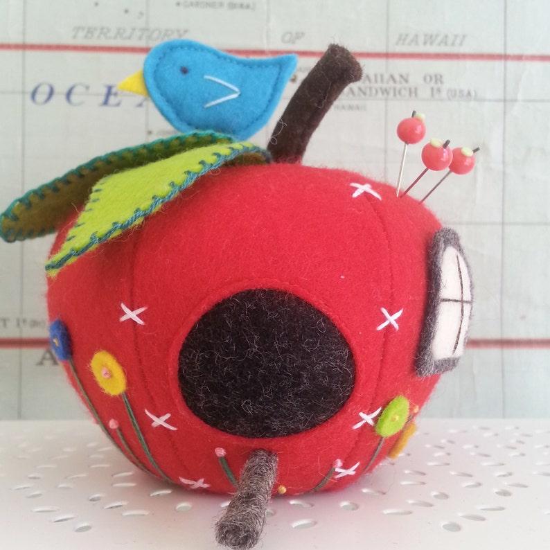 May include: A red felt apple pincushion with a small blue bird on top, a green felt leaf, and a small white window. The apple has a hole in the side for pins and is decorated with colorful felt flowers and stitches.