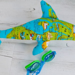 May include: A blue and green plush shark toy with a map design. The shark is lying on a white wooden surface with a pair of blue and green swimming goggles next to it.