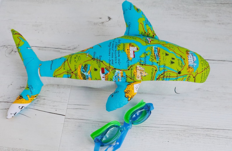 May include: A blue and green plush shark toy with a map design. The shark is lying on a white wooden surface with a pair of blue and green swimming goggles next to it.