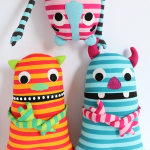 May include: Three striped plush toys with different colours and patterns. The toys have large eyes, a mouth, and horns. The toys are made of soft fabric and are suitable for children.