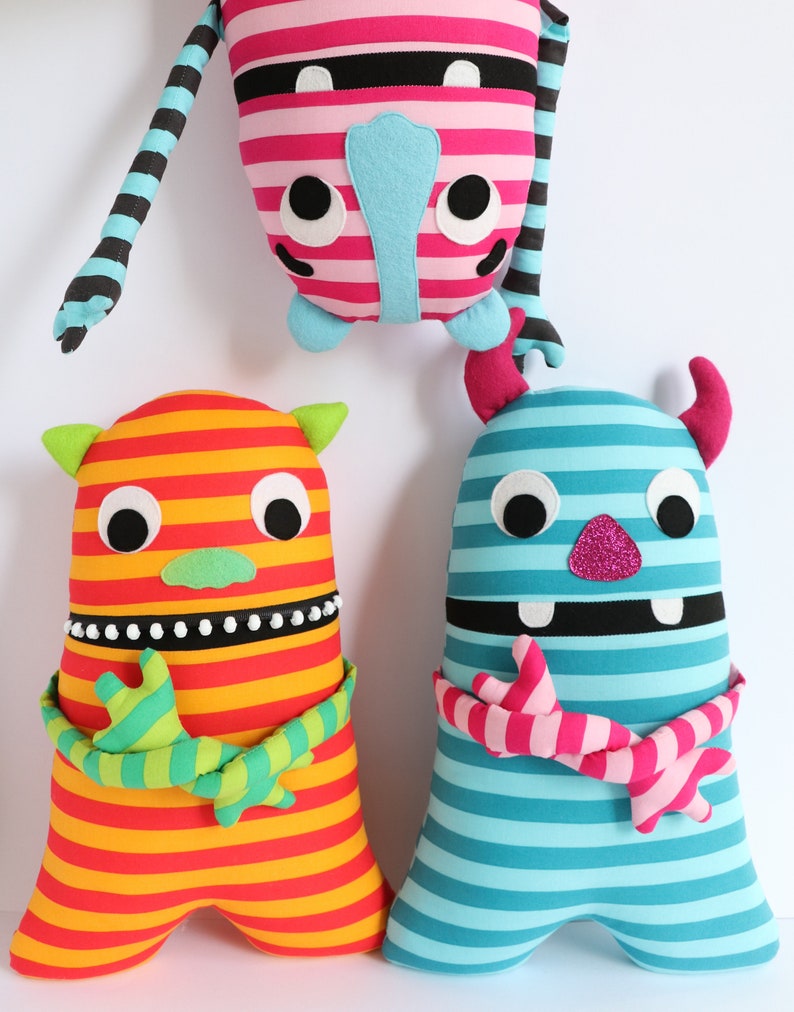May include: Three striped plush toys with different colours and patterns. The toys have large eyes, a mouth, and horns. The toys are made of soft fabric and are suitable for children.