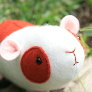 May include: A white and red plush toy shaped like a guinea pig. The toy has a pink nose and black eyes.