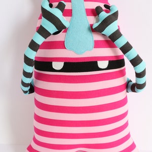May include: A pink and white striped plush toy with blue ears and arms. The arms are striped black and blue. The toy has a black mouth and white teeth. The toy has a blue nose.