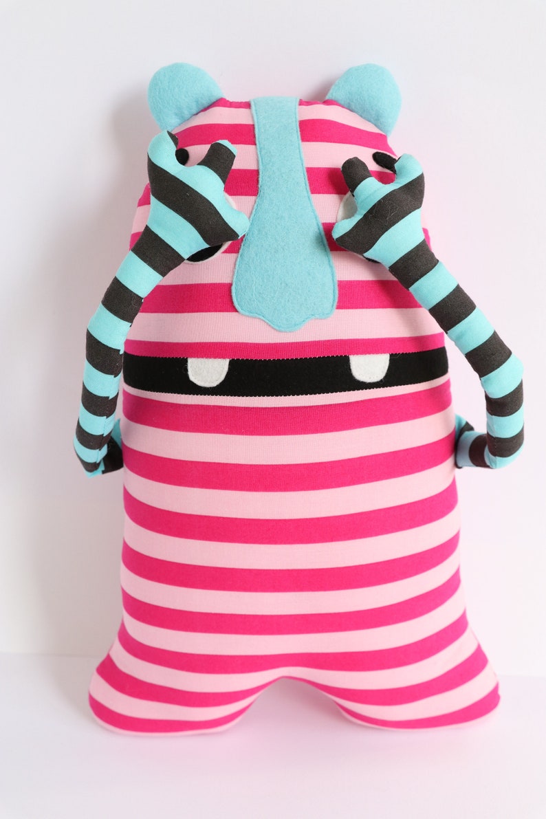 May include: A pink and white striped plush toy with blue ears and arms. The arms are striped black and blue. The toy has a black mouth and white teeth. The toy has a blue nose.