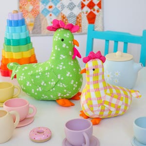 May include: Two fabric chicken plush toys, one green with a white floral pattern and one with a pink and yellow plaid pattern. Both have bright orange feet, beaks, and pink combs. They are surrounded by colourful toy tea cups, a donut, and a stacker.