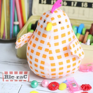 K&ouml;nnte beinhalten: Ein wei&szlig;-orangefarbener Gingham-Stoff-H&uuml;hner-Nadelkissen mit einem rosa Kamm und einem gelben Schnabel. Das Nadelkissen ist umgeben von N&auml;hzubeh&ouml;r, darunter Kn&ouml;pfe, Nadeln und ein Schild mit der Aufschrift "Ric rac".