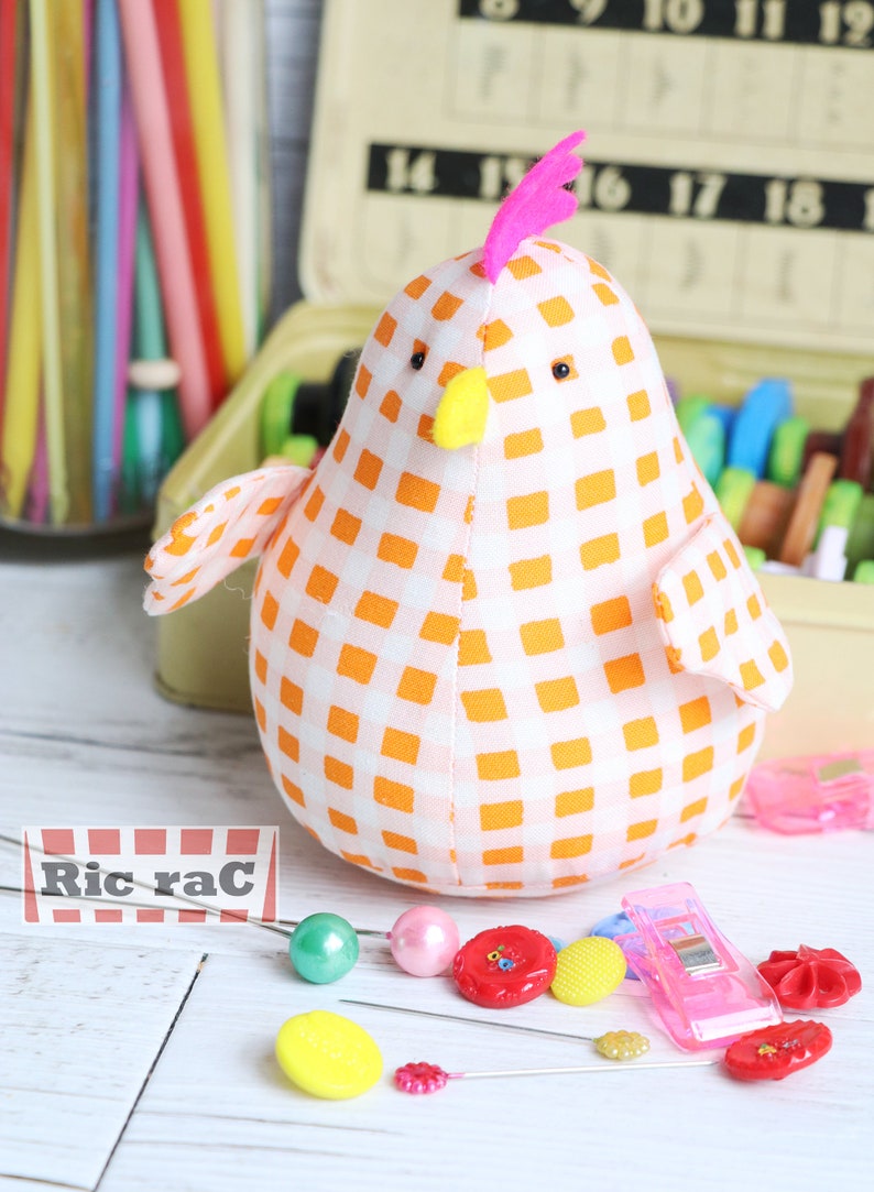 K&ouml;nnte beinhalten: Ein wei&szlig;-orangefarbener Gingham-Stoff-H&uuml;hner-Nadelkissen mit einem rosa Kamm und einem gelben Schnabel. Das Nadelkissen ist umgeben von N&auml;hzubeh&ouml;r, darunter Kn&ouml;pfe, Nadeln und ein Schild mit der Aufschrift "Ric rac".