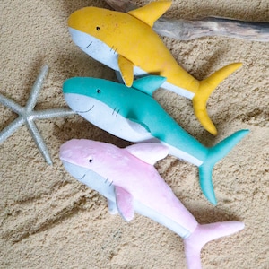 May include: Three plush shark toys on a sandy background. The sharks are yellow, teal, and pink.