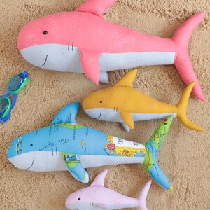 May include: Four plush shark toys on a sandy background. The largest shark is pink and white, the second largest is yellow and white, the third largest is blue and white with a map pattern, and the smallest is pink and white.