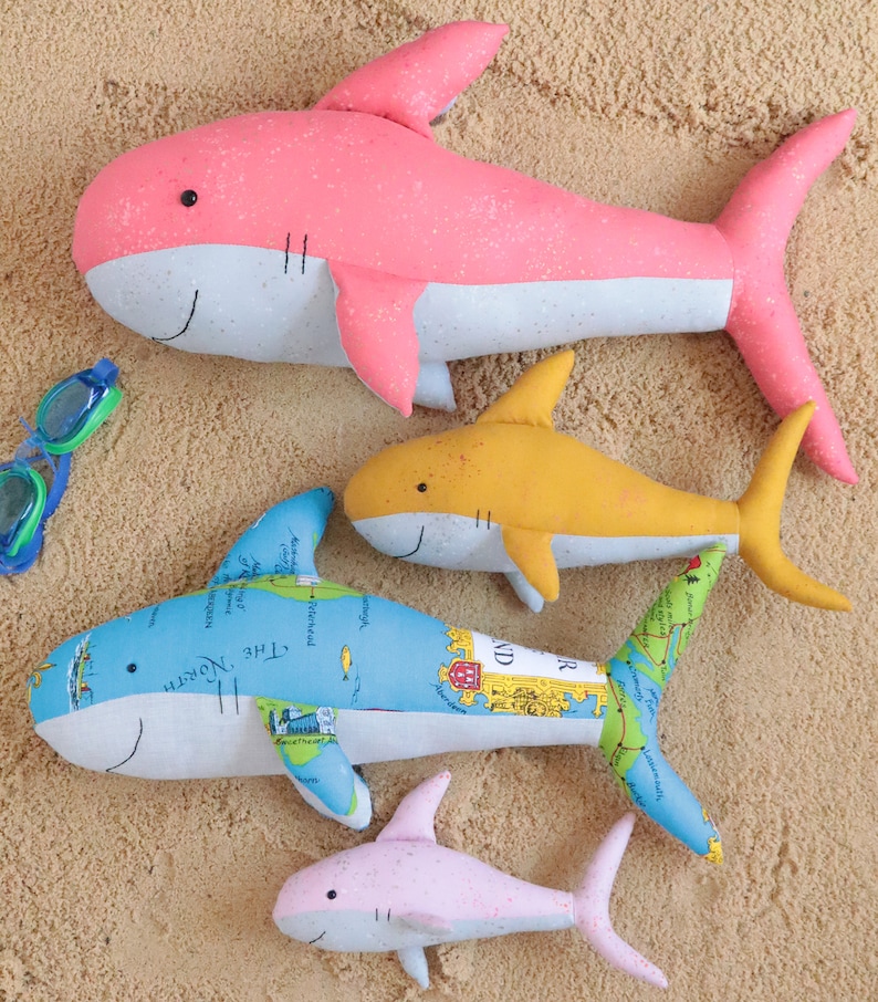 May include: Four plush shark toys on a sandy background. The largest shark is pink and white, the second largest is yellow and white, the third largest is blue and white with a map pattern, and the smallest is pink and white.