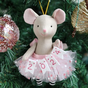 May include: A beige fabric mouse ornament with large ears and a pink stitched nose. The mouse wears a pink tulle skirt with white and pink polka dots and holds a white felt banner with the red embroidered text 2024, featuring black and white striped legs.