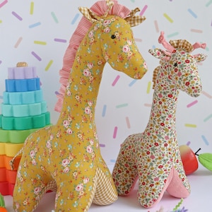 May include: Two stuffed giraffe toys made of floral fabric. The larger giraffe is mustard yellow with pink and white flowers, while the smaller one features a dense multi-colored floral print. Both have gingham accents and pink ruffled manes.