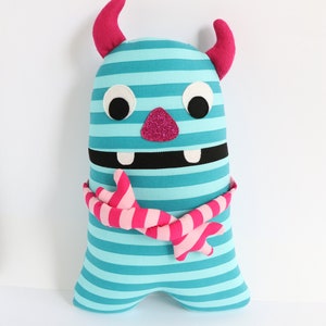 May include: A blue and pink striped monster plush toy with a pink glitter nose, black eyes, and a pink and white striped scarf.