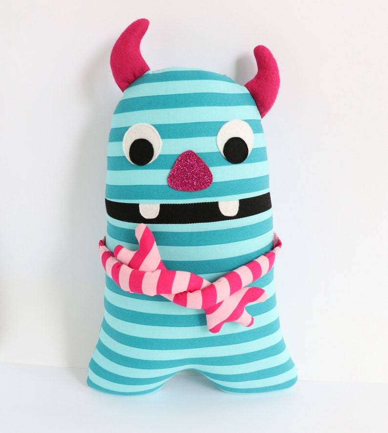 May include: A blue and pink striped monster plush toy with a pink glitter nose, black eyes, and a pink and white striped scarf.