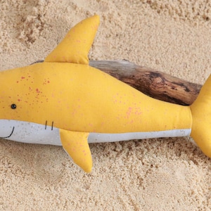 May include: A yellow plush shark toy with pink speckles, lying on a sandy surface with a piece of driftwood.
