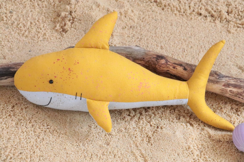 May include: A yellow plush shark toy with pink speckles, lying on a sandy surface with a piece of driftwood.