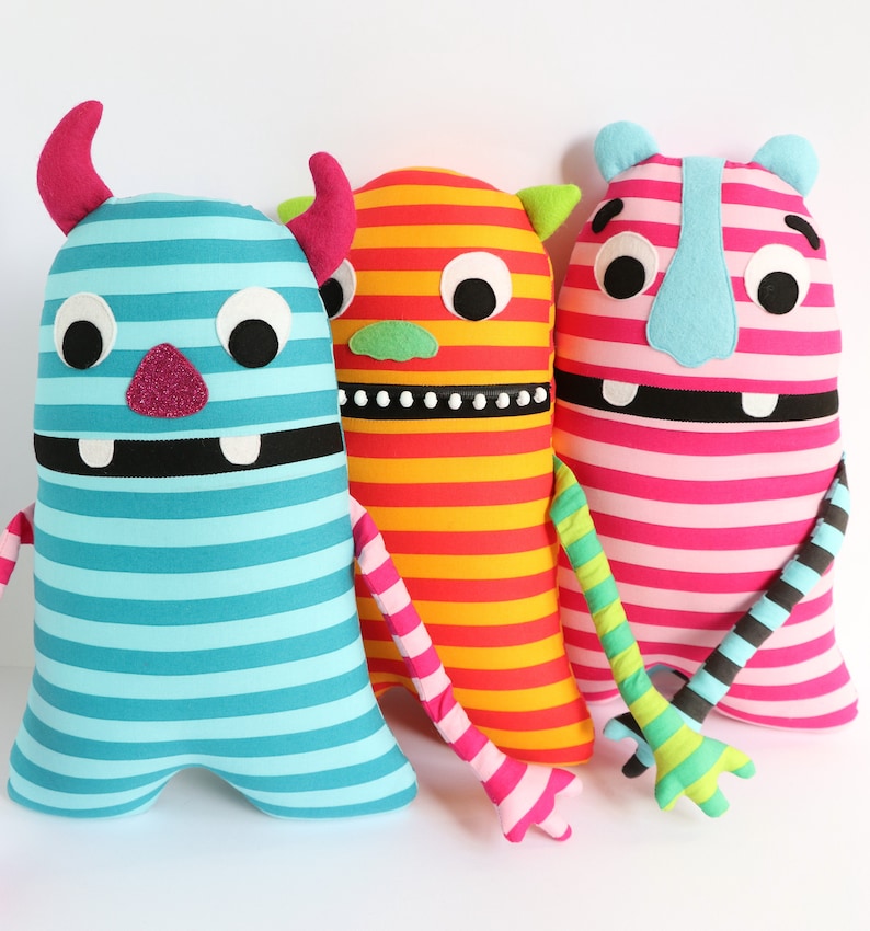 May include: Three striped plush toys shaped like monsters. The monster on the left is blue and turquoise with a black mouth and white eyes. The middle monster is red and yellow with a black mouth and white eyes. The monster on the right is pink and white with a black mouth and white eyes.