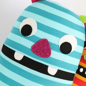 May include: A blue and white striped plush toy monster with pink felt horns, black felt eyes, a pink glitter nose, and a black felt mouth with two white felt teeth.