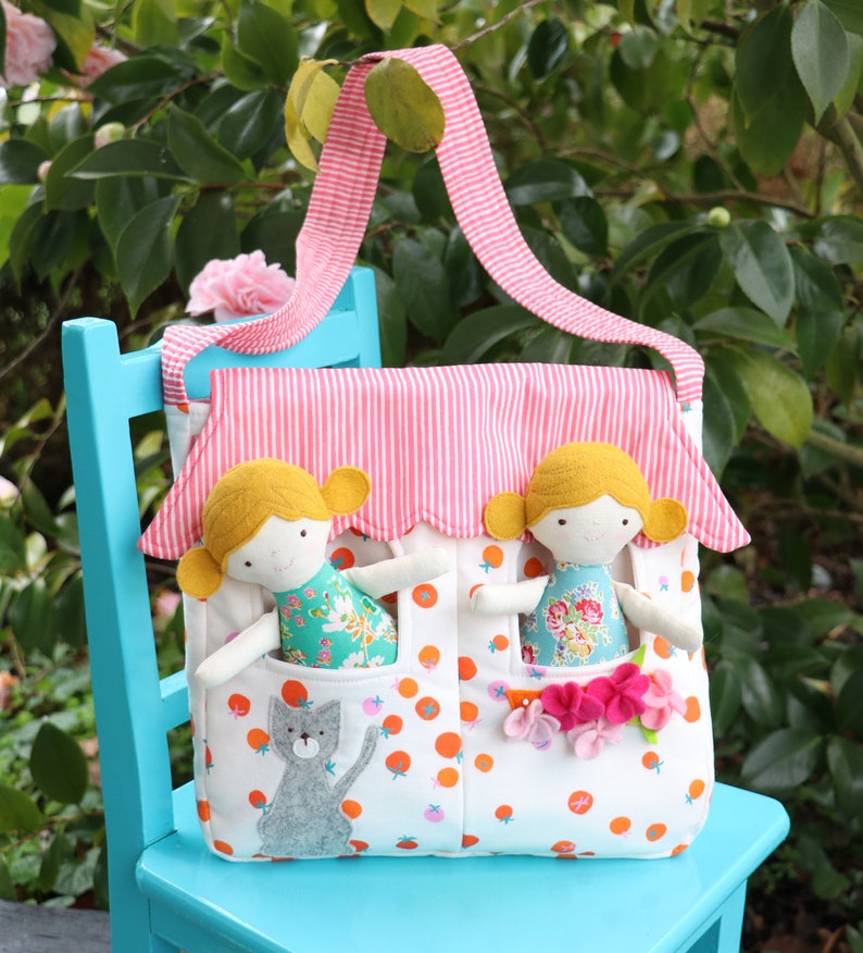 May include: A white and orange fabric tote bag with a pink and white striped roof. The bag has two pockets with a doll in each pocket. The dolls have blonde hair and are wearing colorful dresses. There is a gray cat in the bottom pocket.