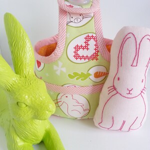 May include: A green bunny figurine sits next to a pink fabric bunny with embroidered details. A green and pink fabric basket with a handle and a pink button is in the background. The basket has a floral pattern with bunnies and hearts.
