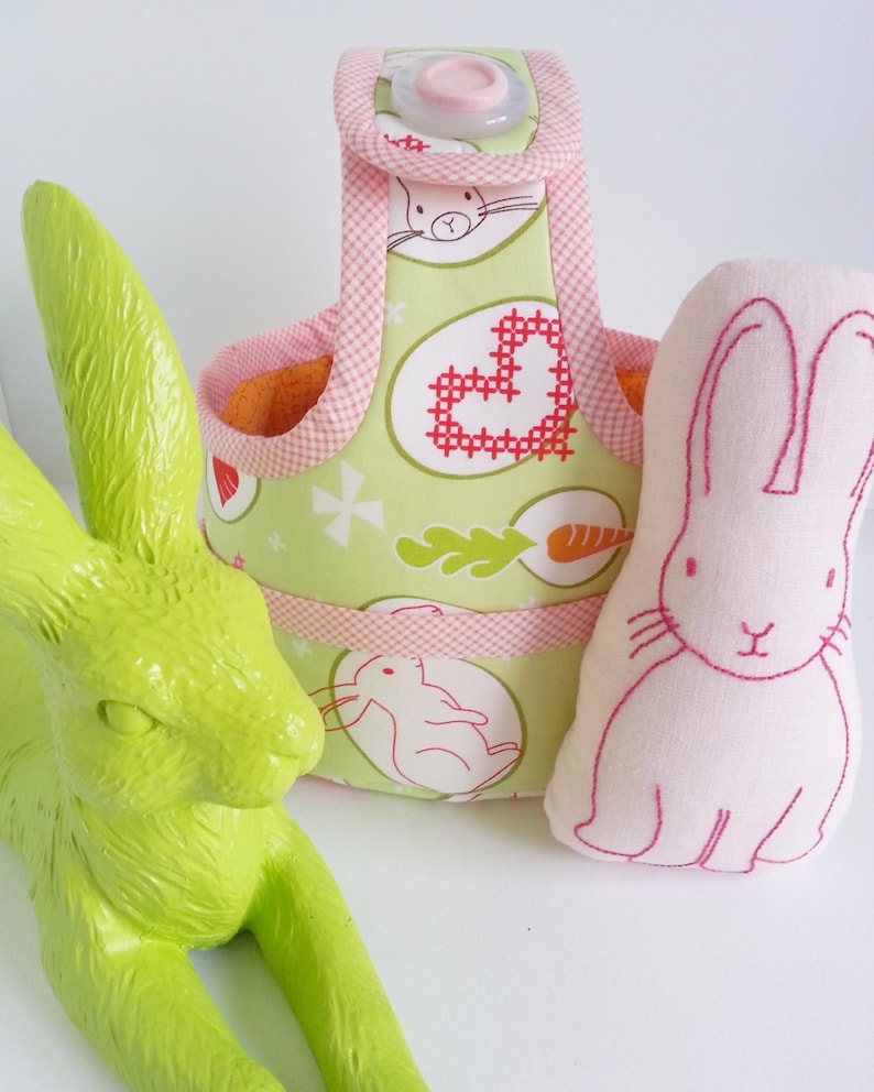 May include: A green bunny figurine sits next to a pink fabric bunny with embroidered details. A green and pink fabric basket with a handle and a pink button is in the background. The basket has a floral pattern with bunnies and hearts.