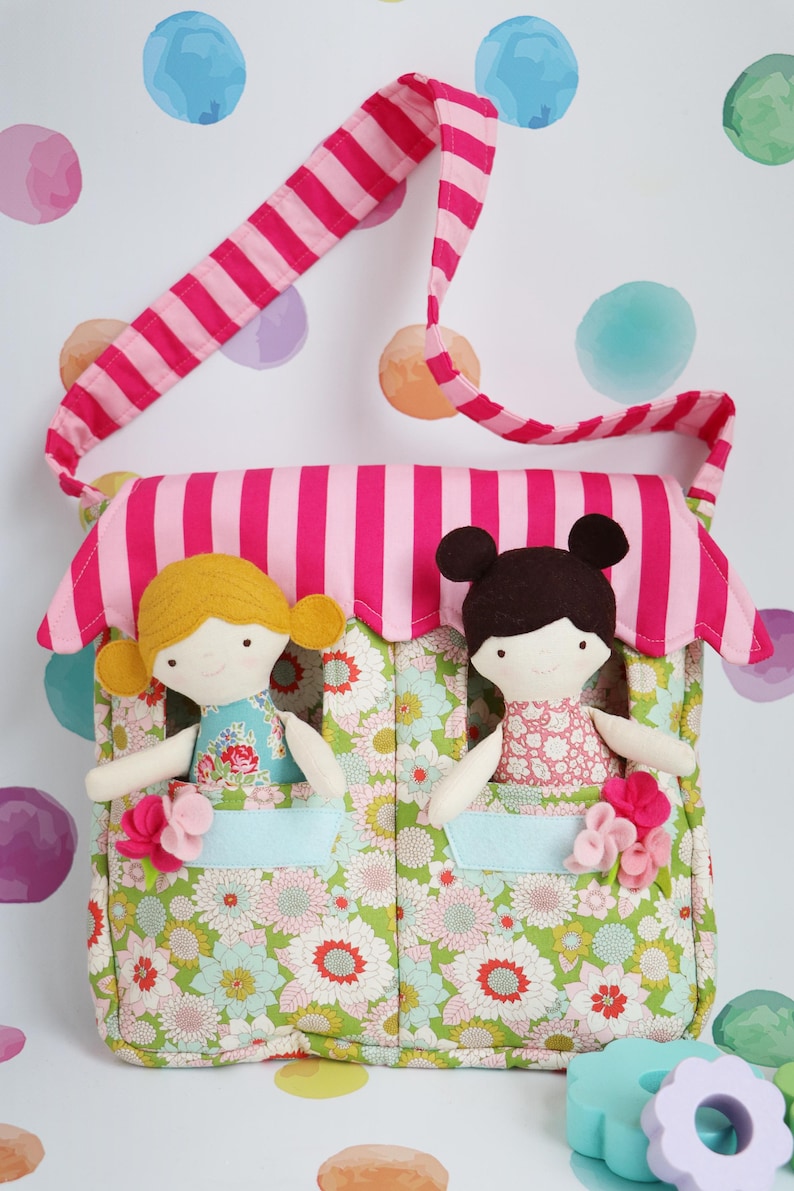 May include: A pink and white striped fabric bag with two felt dolls inside. The dolls have brown and blonde hair and are wearing floral dresses. The bag has a floral pattern and two pockets.