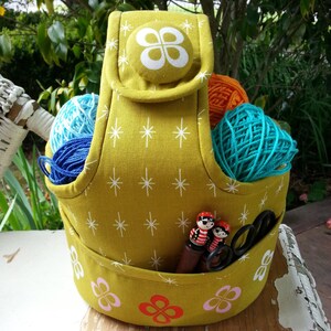 May include: A mustard yellow fabric yarn holder with a white star pattern. The holder has a handle and a pocket for storing yarn and other craft supplies. The holder is filled with colorful yarn balls and knitting needles.