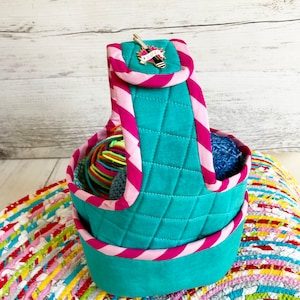 May include: Teal quilted yarn storage basket with a pink and white striped handle. The basket is filled with colorful yarn balls and has a small gold pin with a needle and the word "YARN" on it.