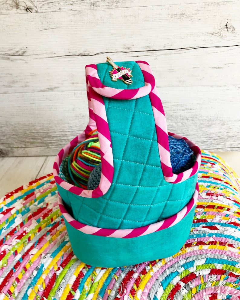 May include: Teal quilted yarn storage basket with a pink and white striped handle. The basket is filled with colorful yarn balls and has a small gold pin with a needle and the word "YARN" on it.
