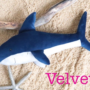 May include: A plush blue and white shark toy with a white belly and a blue tail fin. The toy is lying on a sandy surface with a piece of driftwood and a silver starfish. The word "Velvet" is written in pink letters below the toy.