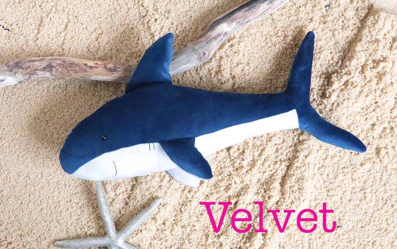 May include: A plush blue and white shark toy with a white belly and a blue tail fin. The toy is lying on a sandy surface with a piece of driftwood and a silver starfish. The word "Velvet" is written in pink letters below the toy.