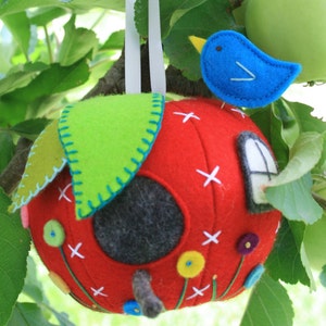 May include: A red felt apple birdhouse with a green felt leaf on top, a blue felt bird, and a small window. The birdhouse is hanging from a tree branch.