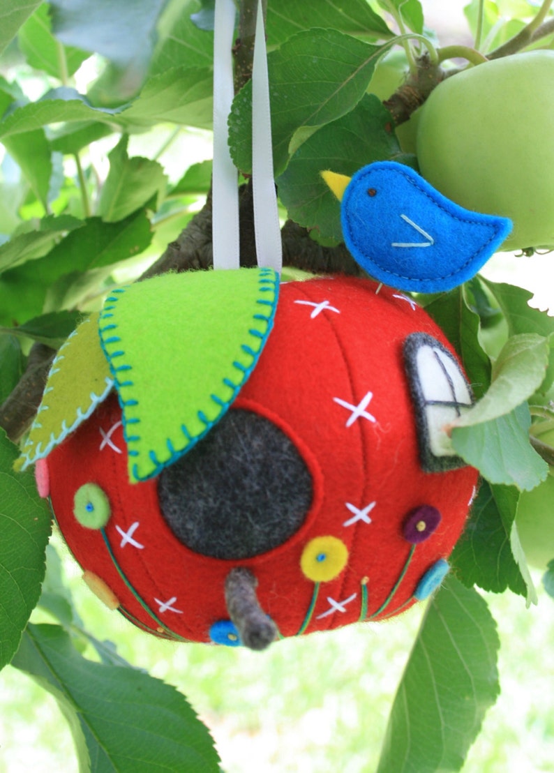 May include: A red felt apple birdhouse with a green felt leaf on top, a blue felt bird, and a small window. The birdhouse is hanging from a tree branch.