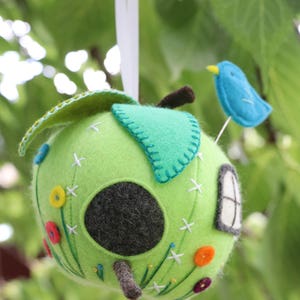 May include: A green felt apple birdhouse with a small blue bird perched on top. The apple has a round hole for the bird to enter and is decorated with embroidered flowers and white stitches.